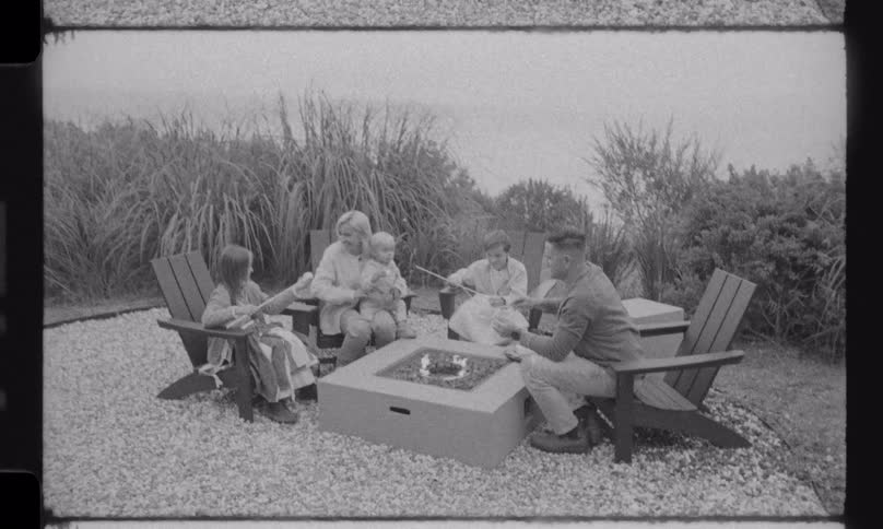 A family gathers around an outdoor fire pit to roast marshmallows in this vintage, black and white styled home movie