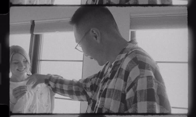 A loving expectant couple smiles while looking at baby clothes together in a grainy black and white vintage-style film