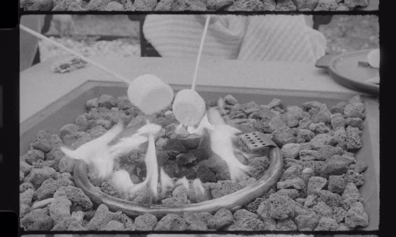 A nostalgic black and white vintage film shows a happy family, including a mother and her sons, roasting marshmallows over a fire pit