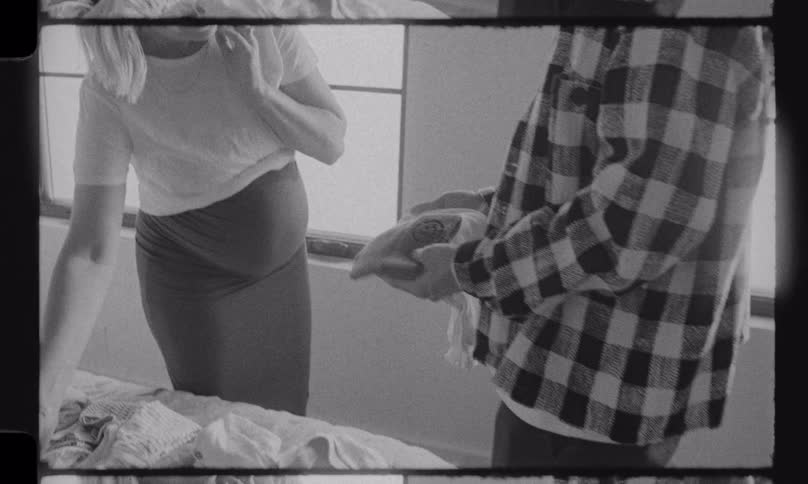 A smiling, expectant couple happily sorts through baby clothes together in their bedroom, captured in a nostalgic black and white film style