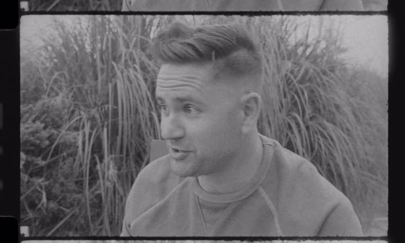 Vintage-style black and white footage captures a father telling an animated story to his family as they roast marshmallows around a backyard fire pit