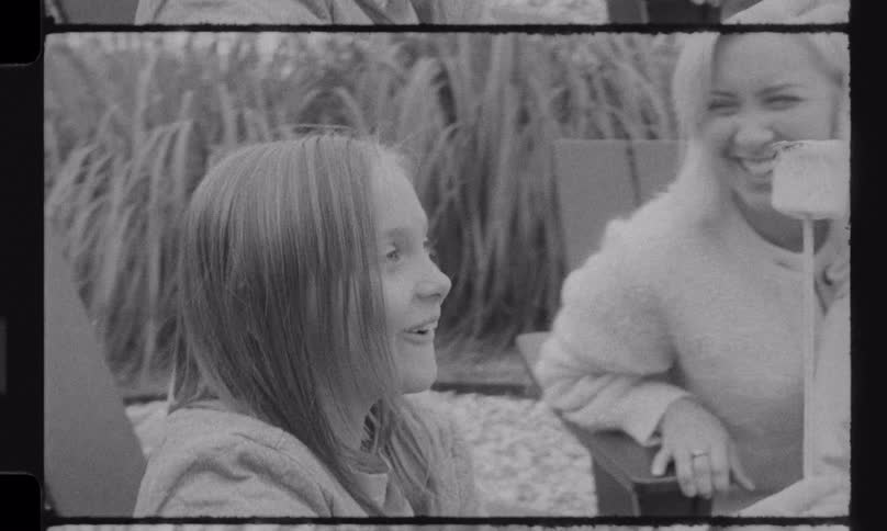 A classic black and white home movie captures a mother and her children joyfully roasting and eating a marshmallow outdoors