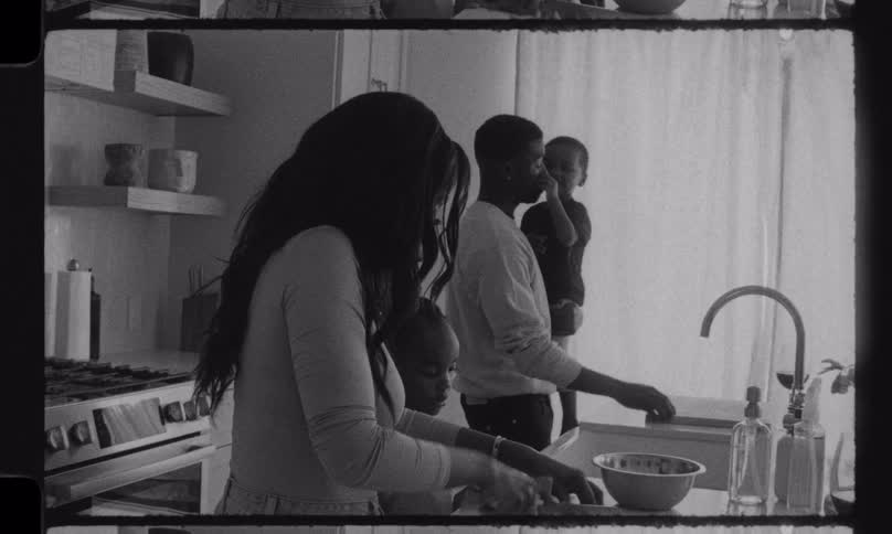 Vintage-style black and white footage captures a tender family moment in the kitchen as a mother prepares a meal