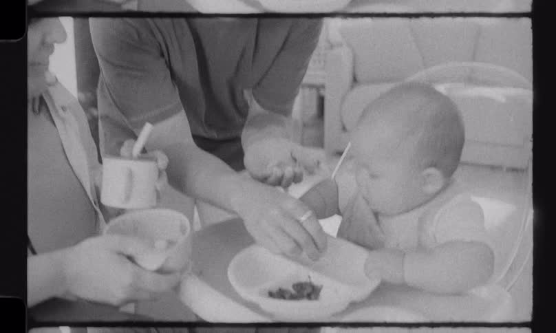 Vintage black and white home movie footage of a mother and father feeding their baby in a high chair at the table