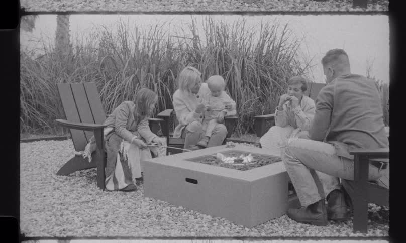 Vintage-style black and white footage captures a happy family roasting marshmallows and making s'mores together around a modern fire pit in their backyard