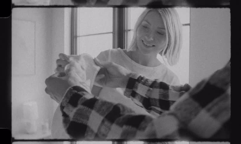 An expectant couple happily sorts through baby clothes in their bedroom in a nostalgic black and white home movie