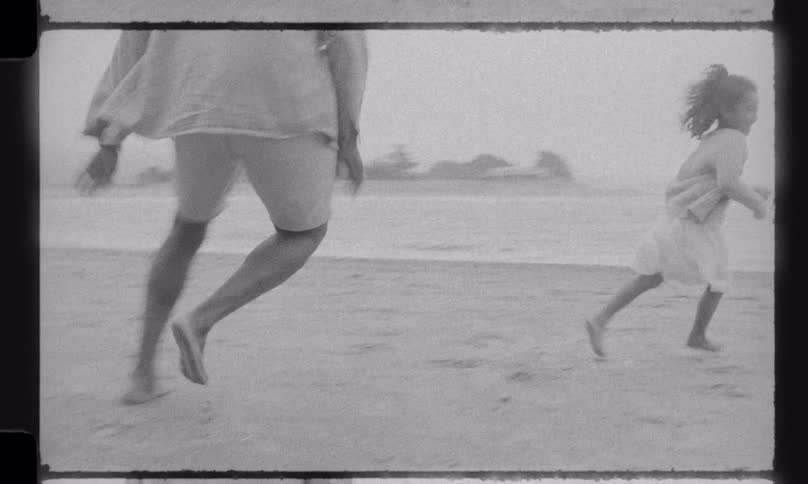 A father playfully chases his laughing daughter on a sandy beach, catching her in a warm embrace in this nostalgic, vintage-style footage