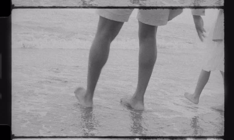 A father and his young daughter enjoy a walk on the beach, with waves washing over their bare feet in this nostalgic, vintage-style footage