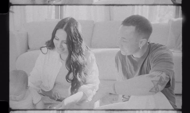 Vintage-style black and white footage shows a happy young family playing together with their baby on the living room floor