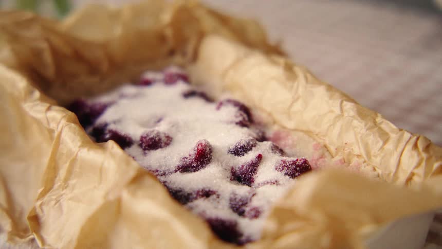 A close-up of white sugar being poured over red berries in a parchment-lined baking dish