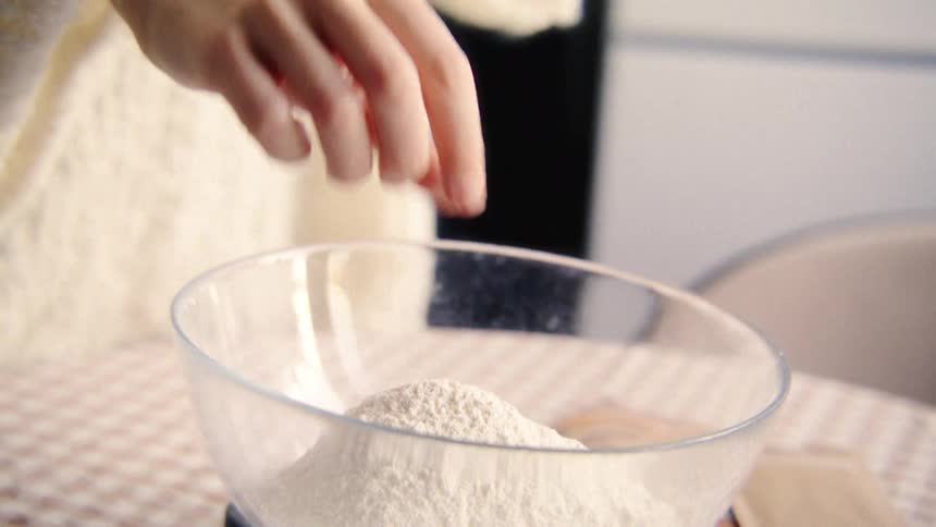 Slow motion close-up of a baker sprinkling ingredients into a glass bowl of flour