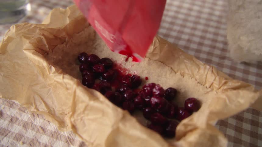 A close-up, slow-motion shot shows red syrup being poured from a packet onto cherries in a baking paper-lined dish