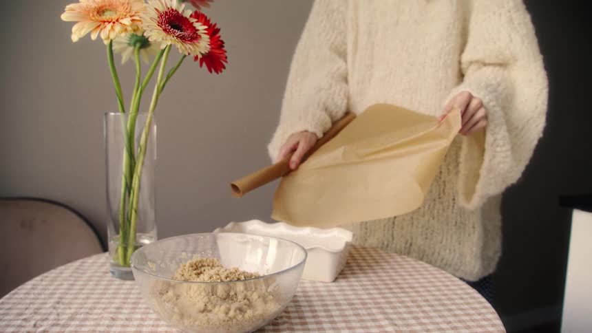 A woman in a cozy sweater unrolls a sheet of parchment paper over a table with baking ingredients and a vase of flowers