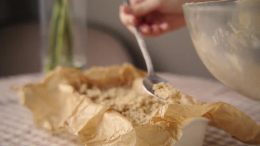 A person carefully spoons a crumble mixture from a glass bowl into a baking tin lined with parchment paper in slow motion
