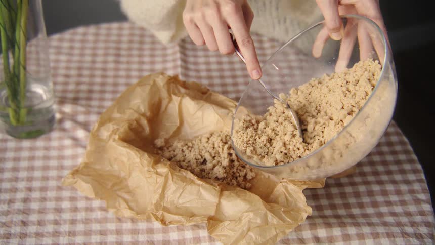 A person in a cozy sweater carefully transfers crumble topping from a glass bowl to a parchment-lined baking dish