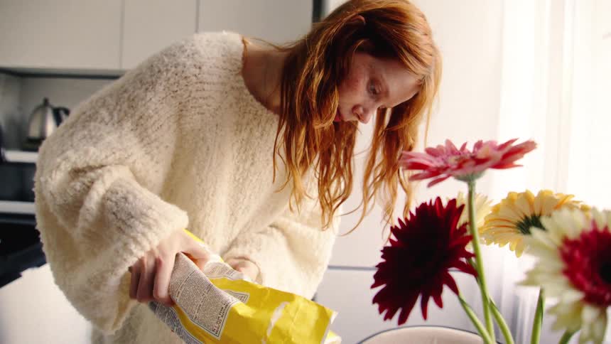 A young woman with red hair in a cozy sweater carefully pours flour into a bowl to bake in her sunlit kitchen