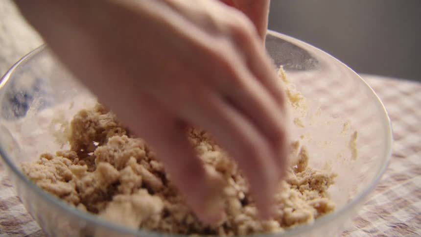 Slow motion closeup of a person's hands kneading and mixing a crumble dough in a clear glass bowl