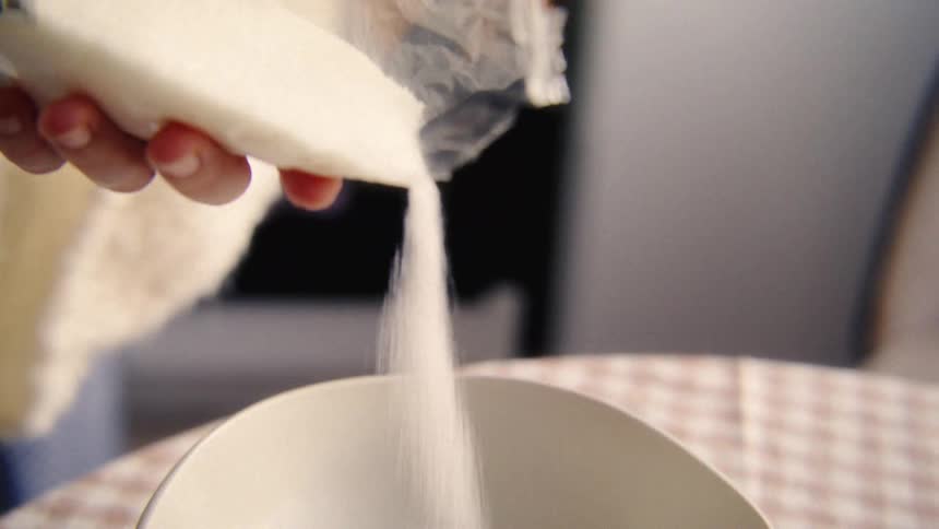 A close-up slow-motion shot of white sugar being poured from a bag into a bowl with a raw egg