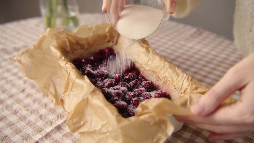 A baker gently sprinkles sugar over ripe cherries in a parchment-lined baking pan in slow motion