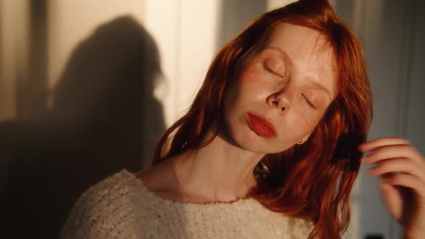 A pensive red-haired woman with freckles runs her hand through her hair in slow motion, illuminated by the warm light of the golden hour