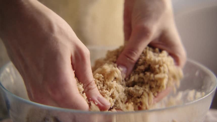 A close-up, slow-motion shot of a baker's hands rubbing together ingredients to make a crumble topping in a glass bowl