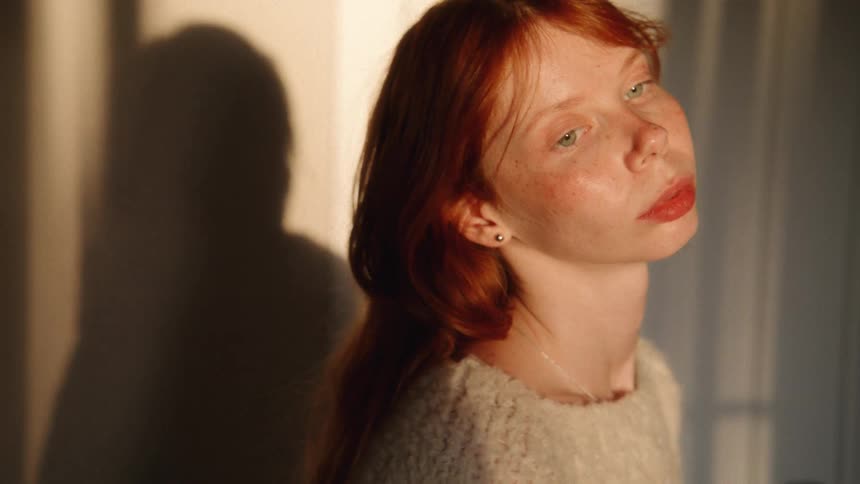 A pensive red-haired woman with freckles basks in the golden hour sunlight, her shadow cast on the wall behind her