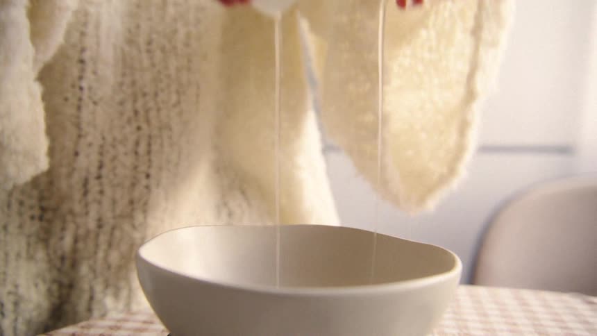 A close-up slow-motion shot shows a person's hands carefully cracking a fresh egg into a white bowl