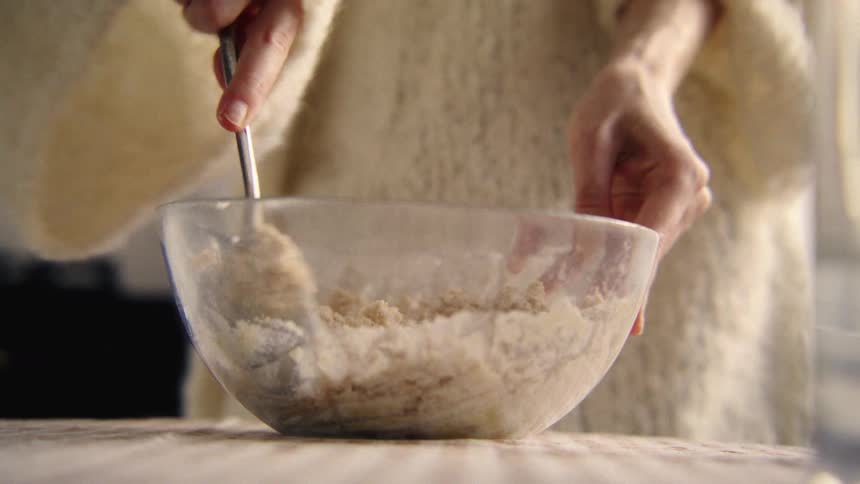 A person in a cozy white sweater slowly mixes baking ingredients with a fork in a clear glass bowl