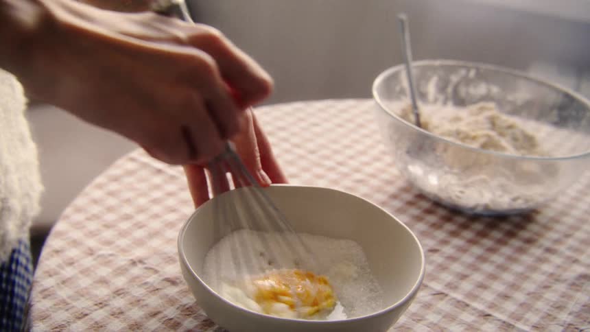 Slow motion close-up of hands whisking an egg and sugar in a white bowl to prepare a cake batter