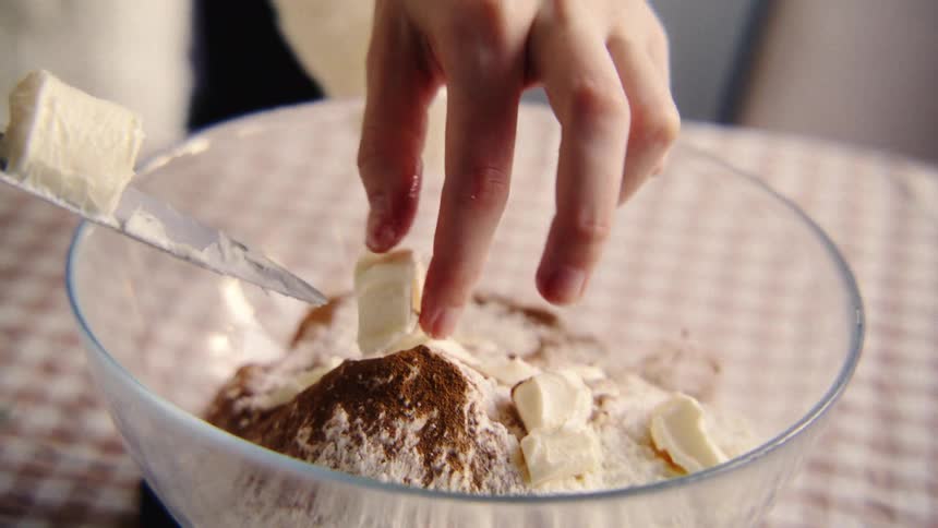 A close-up slow-motion shot shows a baker adding cubes of butter to flour and cocoa powder in a glass bowl