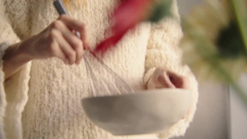 Close-up of a young redhead wearing a cozy sweater whisking ingredients in a bowl in a bright kitchen
