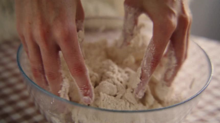 A closeup slow-motion shot of a baker's hands kneading flour and butter in a glass bowl to make dough