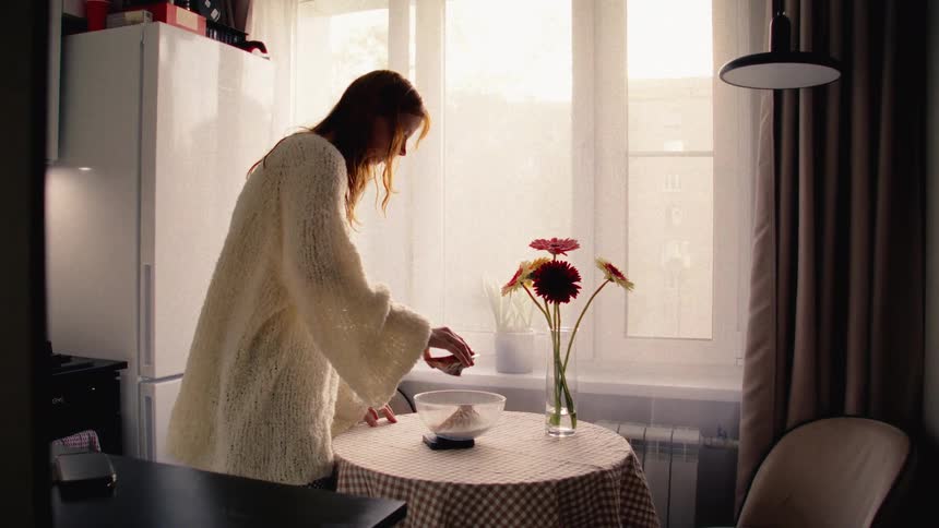 A woman in a cozy sweater carefully cracks an egg into a bowl of flour on a kitchen scale in a sunlit kitchen
