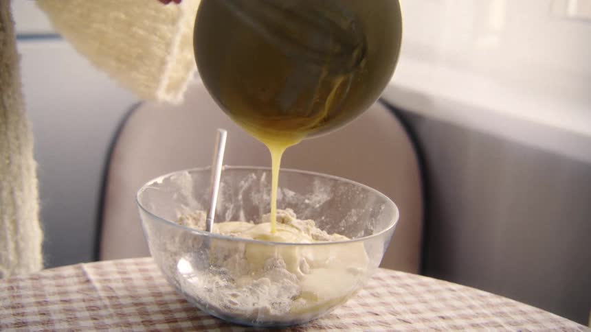 A baker in a cozy sweater pours a whisked egg mixture into a glass bowl with flour in slow motion