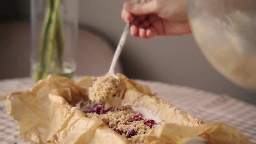 A baker slowly sprinkles a crumbly streusel topping over a berry mixture in a parchment-lined baking pan