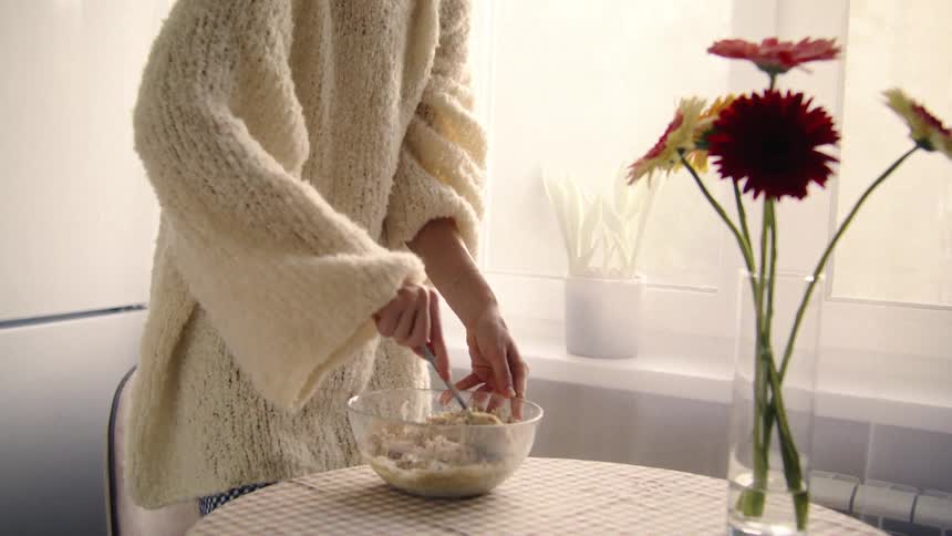 A person in a cozy white sweater mixes ingredients in a glass bowl in a brightly lit kitchen