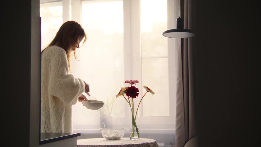 A woman in a cozy sweater whisks ingredients in a bowl while standing in a softly lit kitchen by a window