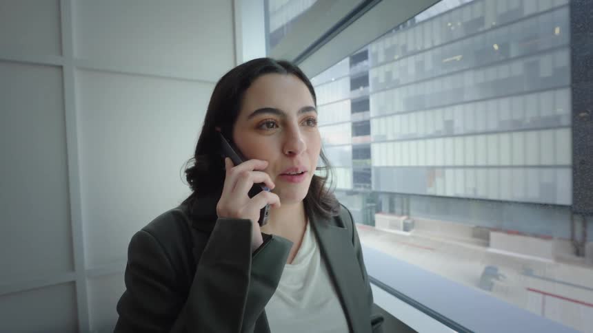 A young businesswoman smiles while having a pleasant phone conversation by the window in a modern high-rise building