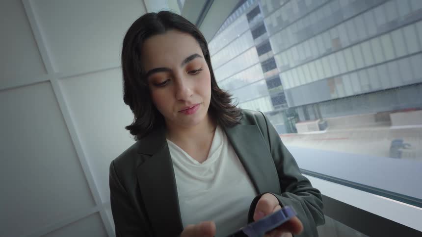 A young businesswoman smiles as she texts on her smartphone while standing by a large window in a modern office building