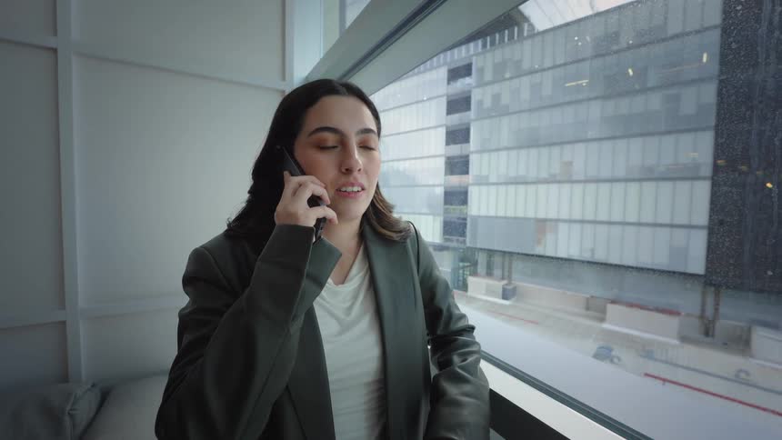 A professional young businesswoman in a blazer talks on her phone while standing by a large window in a modern office