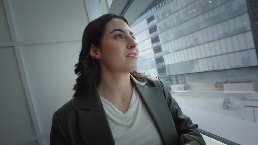 A young businesswoman stops using her phone to look thoughtfully out a large window in a modern high-rise office, smiling with hope