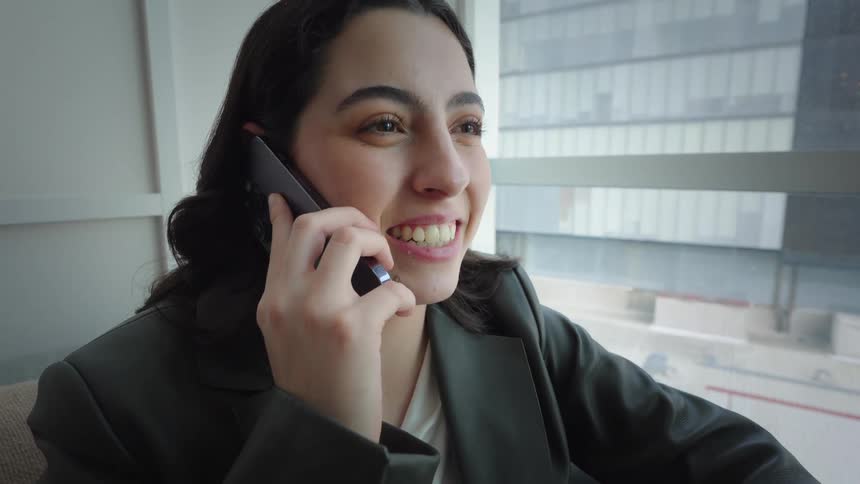 A smiling businesswoman in a suit talks on her smartphone while sitting on a couch in a modern office with a large window