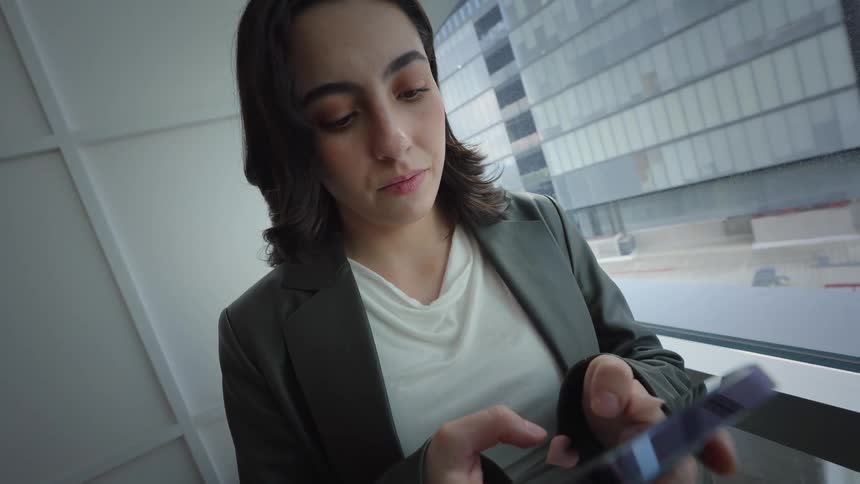 A young businesswoman in a blazer stands by a large office window, intently focused on scrolling through her smartphone as the camera slowly pushes in