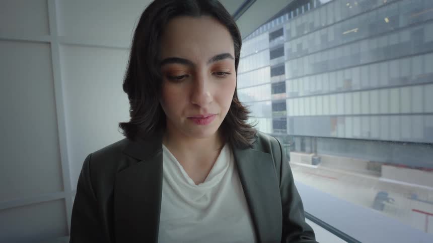 Push-in shot of a young businesswoman focused on her smartphone while standing by a large window in a modern office