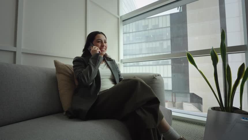 A relaxed professional woman smiles while talking on her smartphone, sitting on a couch by a large window overlooking the city