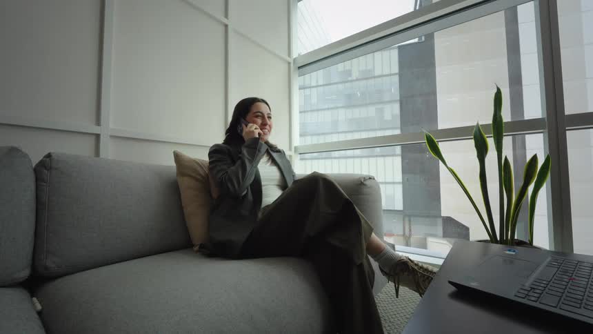 A young businesswoman smiles while talking on her phone, relaxing on a couch in a modern high-rise office