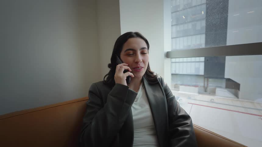 A smiling businesswoman answers a phone call while relaxing on a leather sofa in a modern office with a city view