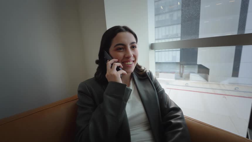 A young, stylish businesswoman relaxes on a couch and answers her phone, her face lighting up with a smile during the conversation