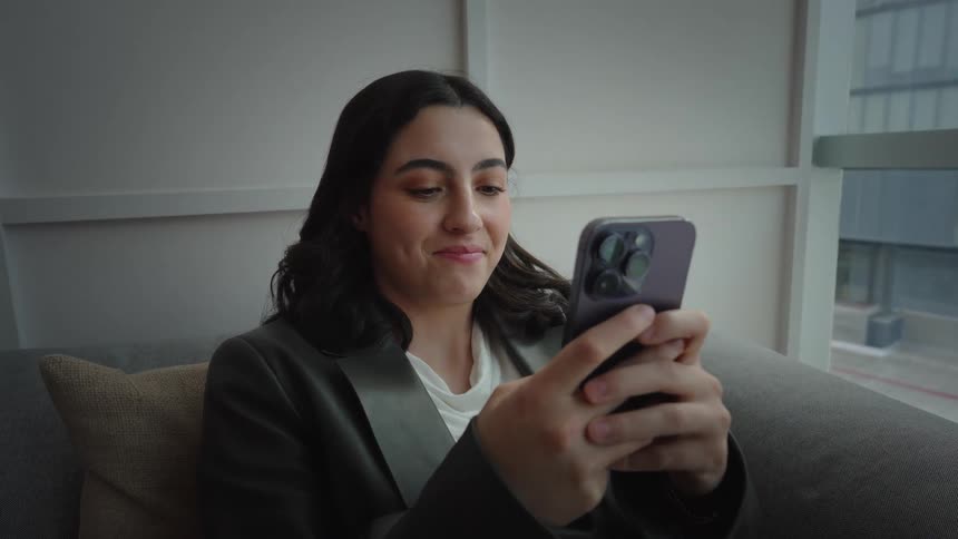 A young businesswoman smiles while using her smartphone as she sits on a couch in a modern office