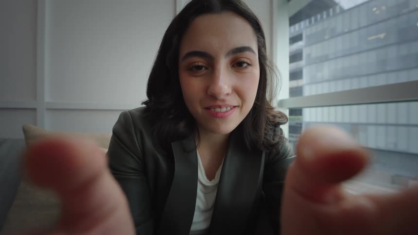 First-person perspective of a young woman smiling during a video call, her fingers framing the screen in a modern city office